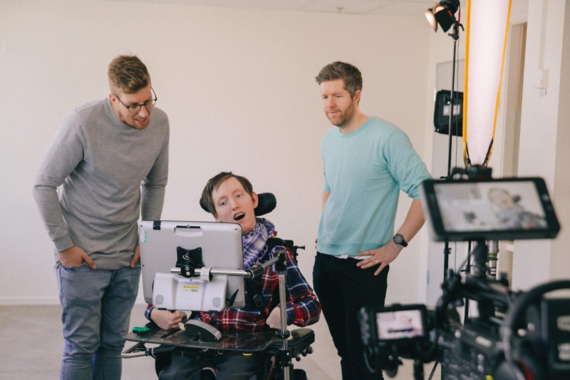 A disabled man in a motorised wheelchair using TV production equipment. There are 2 other men standing either side of him.